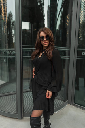 Fashionable beautiful glamor stylish street woman with cool sunglasses in a trendy fashion black dress stands near a modern building in the city. Pretty ladyの写真素材