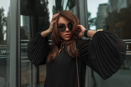 Fashion female street portrait of beautiful stylish glamorous cool woman with vintage sunglasses in fashionable black dress near modern glass building. Pretty ladyの写真素材