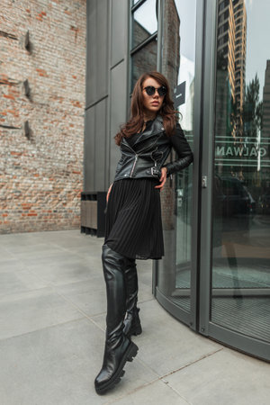 Cool stylish beautiful chic model girl with fancy sunglasses in fashion black street rock outfit with leather jacket, dress and high shoes standing near a modern building in the cityの写真素材