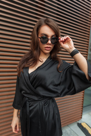 Beautiful fashion fresh brunette woman with hairstyle in a fashionable elegant black dress puts on cool sunglasses and walks in the city near the metal wallの写真素材