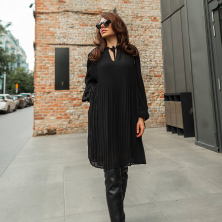 Fashionable beautiful elegant woman model with sunglasses in a fashion black dress with high shoes walks in the city near a brick vintage buildingの写真素材