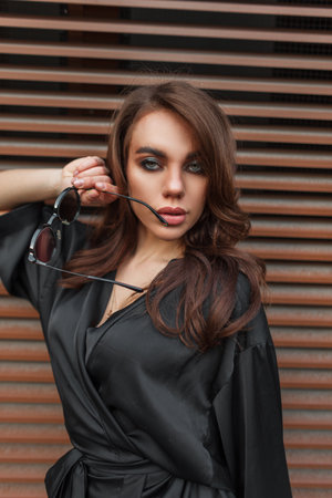 Beautiful elegant stylish glamorous woman with make-up in a fashion black dress with sunglasses stands on the street near a rusty metal wallの写真素材