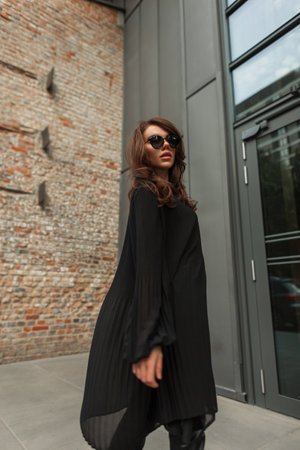 Fashion beautiful woman model with brunet hairstyle with sunglasses in black stylish dress walks on the streetの写真素材