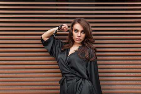 Cool pretty young stylish chic beauty woman with make-up with sunglasses in a black fashion dress stands and poses near a rusty metal wall in the streetの写真素材