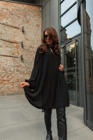 Beautiful fashionable young woman model with cool sunglasses in fashion trendy black clothes with black dress and high leather shoes in the city. Chic pretty glamor ladyの写真素材