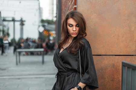 Elegant beauty woman brunette model with hairstyle and makeup in black dress stands near a metal rustic wall in the cityの写真素材