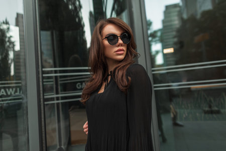 Elegant beautiful young fashionable gorgeous girl model with brunette hairstyle with cool vintage sunglasses in black trendy fashion dress in the cityの写真素材