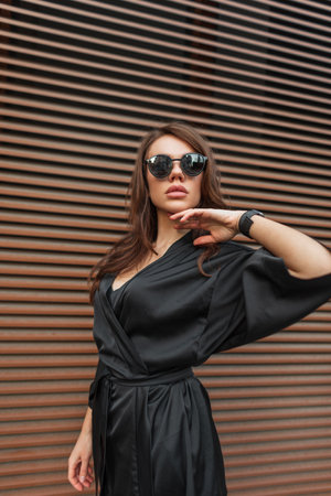 Beautiful fashion young glamor chic model girl with cool sunglasses in a stylish black dress stands and poses near a rusty metal wallの写真素材