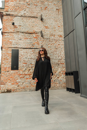 Elegance trendy fashion beauty woman with sunglasses in black fashionable street outfit with dress and high shoes walks in the city near a vintage brick buildingの写真素材
