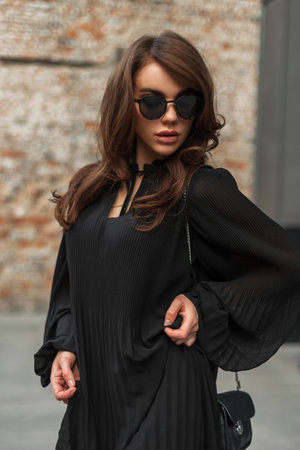 Glamorous beautiful young stylish woman model with vintage sunglasses in a fashion trendy black dress with a small handbag in the city near the brick buildingの写真素材