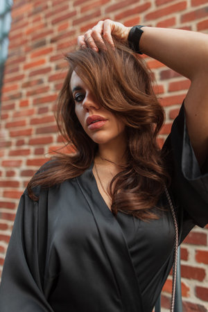 Female stylish portrait of a beautiful fashion woman model with makeup and brunette hair in a trendy black dress on the street near a red brick wallの写真素材