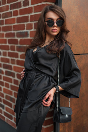 Beautiful fashion young brunette woman model with stylish sunglasses in a trendy fashionable elegant evening suit with a leather black small handbag stands near a vintage brick metal wall in the cityの写真素材