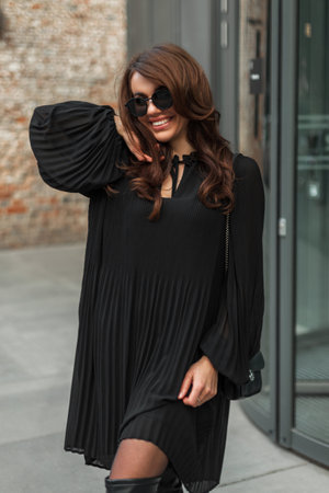 Happy beautiful glamor woman model with hairstyle with stylish vintage sunglasses in a fashion black dress with a handbag walks and has fun in the city. Pretty ladyの写真素材