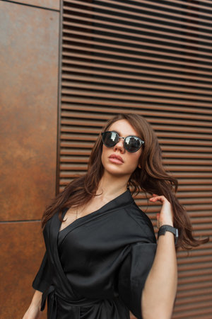 Cool trendy stylish model woman with brunette hair with sunglasses in a fashionable black elegant dress walking outside near a rusty metal wallの写真素材