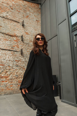 Gorgeous fashion beautiful young lady model with brunette hairstyle with stylish sunglasses in fashionable black dress walks and poses in the city near the building. Pretty glamor woman in outfitの写真素材