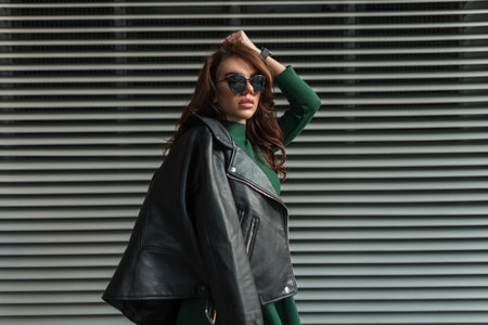 Cool trendy beauty young stylish woman model with fashion sunglasses in green dress with leather black jacket posing on the street near metal lines wallの写真素材