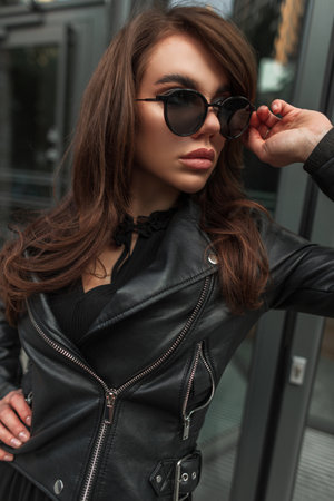Fashion pretty chic woman with brunette hairstyle in black stylish leather jacket and dress wears a cool vintage sunglasses on the street. beauty urban girlの写真素材