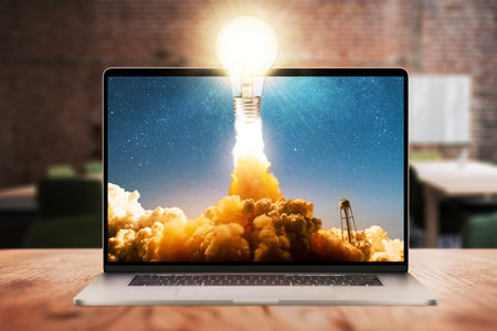 Concept light bulb glows and successfully lift off with smoke and clouds from the laptop display on wooden desktop in office. Successful start up, creative thinking and thinking differentlyの写真素材