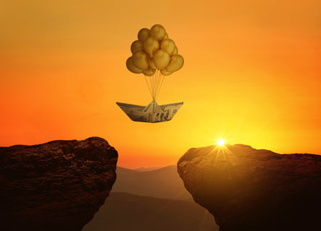 Paper boat dollars with balloons flies over a cliff with rocks at sunset. Economy, business, success and startup, concept. Deposits and insurance, creative idea. World financial crisisの写真素材
