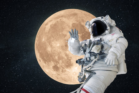 Space man astronaut greets and waves his hand in space on a background of amazing full moon. Space mission wallpaper, concept. Welcome to space. Moon missionの写真素材