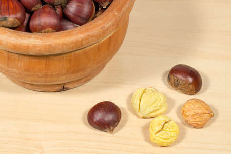 Several peeled chestnuts, one of them with pellicle, beside a wooden bowl full of chestnutsの写真素材