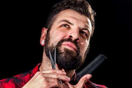 Frontal view of young man trimming his beard with comb and scissors