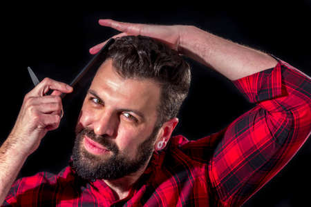 Bearded man with red plaid shirt combs his hair, on blackの写真素材