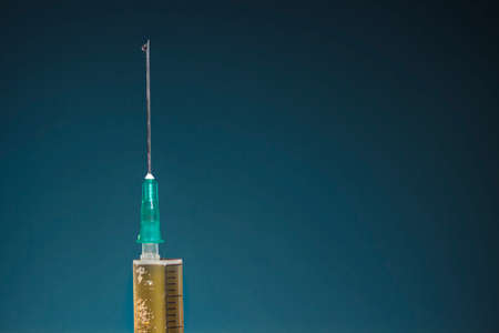 Hypodermic needle with a drop at its tip and the top of syringe with dark yellow drug, on dark blue...