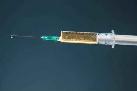 Hypodermic needle with drop at its tip and syringe with dark yellow drug, on dark blue background