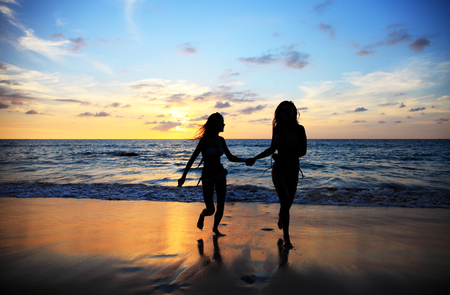 Two young happy women walking on tropical sea beach at sunsetの写真素材