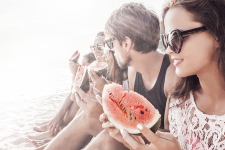 Group of young people sitting on beach and eating watermelonの写真素材