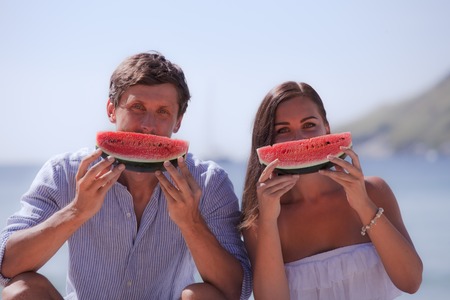 Cheerful couple eating watermelon at sea beachの写真素材