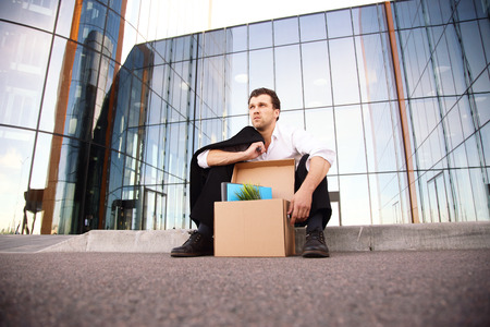Fired business man sitting frustrated and upset on the street near office building with box of his belongings. He lost workの写真素材