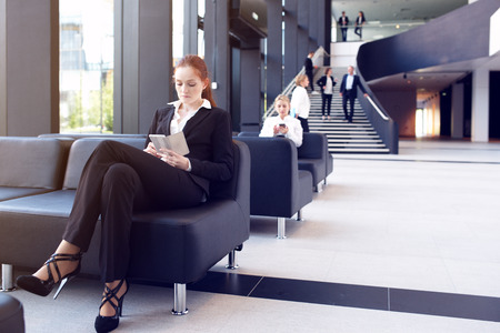 Young businesswoman sitting in lobby and surfing the net with mobile phoneの写真素材