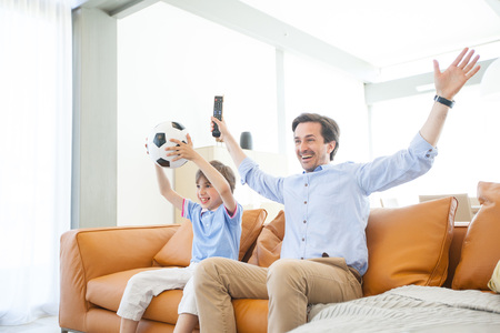 Portrait of happy boy watching soccer match with father on sofa at homeの写真素材