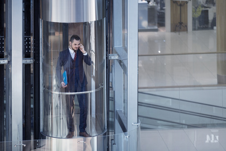 Businessman in modern glass elevator talking by the phone and holding papersの写真素材