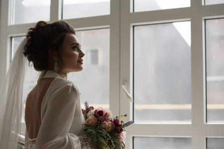 Beautiful bride in her wedding day with flower bouquet looking at windowの写真素材