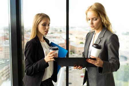 Business women discuss documents and drink coffe in office in front of panoramic windows with view at cityの写真素材