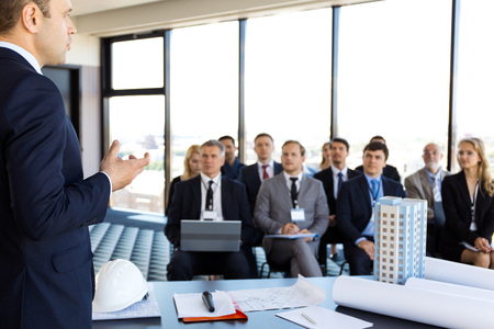 Audience of business people and investors sitting at presentation room, conference hall, view from speaker table with blueprints, model of modern multi storey residential buildingの写真素材