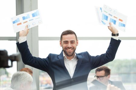 Young joyful businessman standing with arms raised holding data statistics diagrams and smiling , team of his colleagues sitting at the table in the backgroundの写真素材