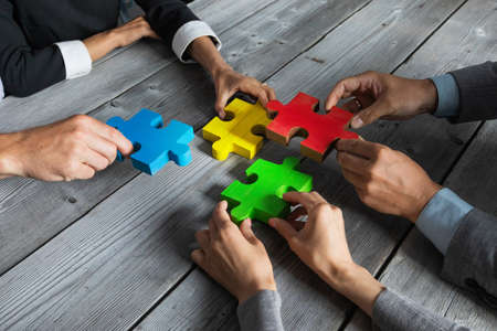 Business people team sitting around meeting table and assembling color jigsaw puzzle pieces unity...