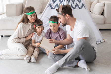 Happy family of parents and two children playing indian at home, wigwam tent, feather roach, reading book about native american indian cultureの写真素材