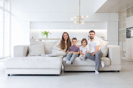Family portrait of young parents with their two children at home, white modern desgin of living roomの写真素材