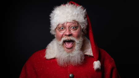 Excited elderly man with white beard wearing santa claus costume looking at camera with big eyes and open mouth, has breaking newsの写真素材