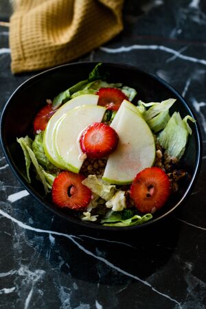 Apple Salad with Strawberry Slices, Buckwheat and Green Leaves in Black Bowl on Dark Granite Surface. Organic Fresh Food.の写真素材
