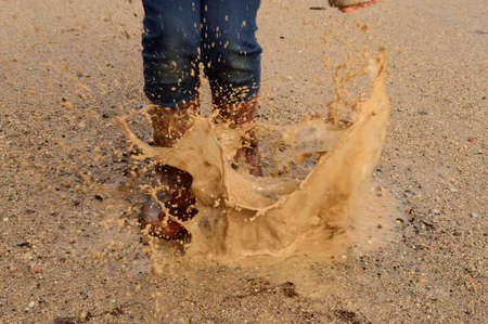 A youngster jumps and splashes into a mud puddle wearing rubber boots.