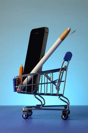 A miniature shopping cart with a cell phone and writing tools inside over a blue background.の写真素材