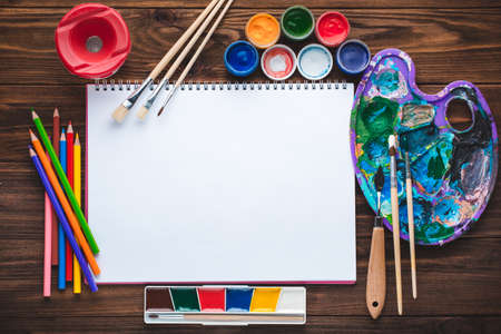 Set of paints, pencils, tools for painting and blank white paper sheet of sketchbook on vintage wooden background. Top view.の写真素材