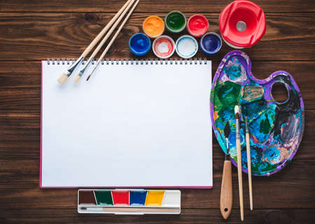 Set of paints, pencils, tools for painting and blank white paper sheet of sketchbook on vintage wooden background. Top view.の写真素材