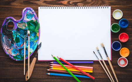 Set of paints, pencils, tools for painting and blank white paper sheet of sketchbook on vintage wooden background. Top view.の写真素材